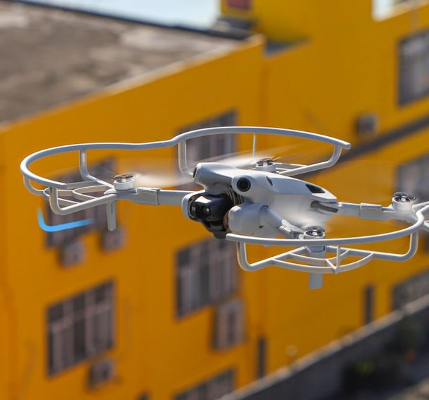 DJI Mini 4 Pro drone with propeller guards flying against a bright yellow building backdrop.